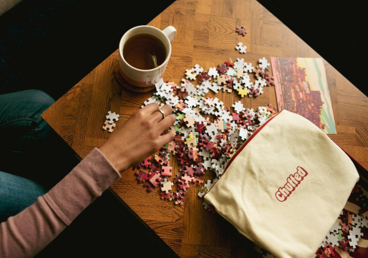 Person working on a puzzle with a mug of tea and a bag labeled 'Chilled' on a wooden table.
