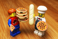 Two LEGO figures on a wooden surface, one holding a pizza and the other a loaf of bread.